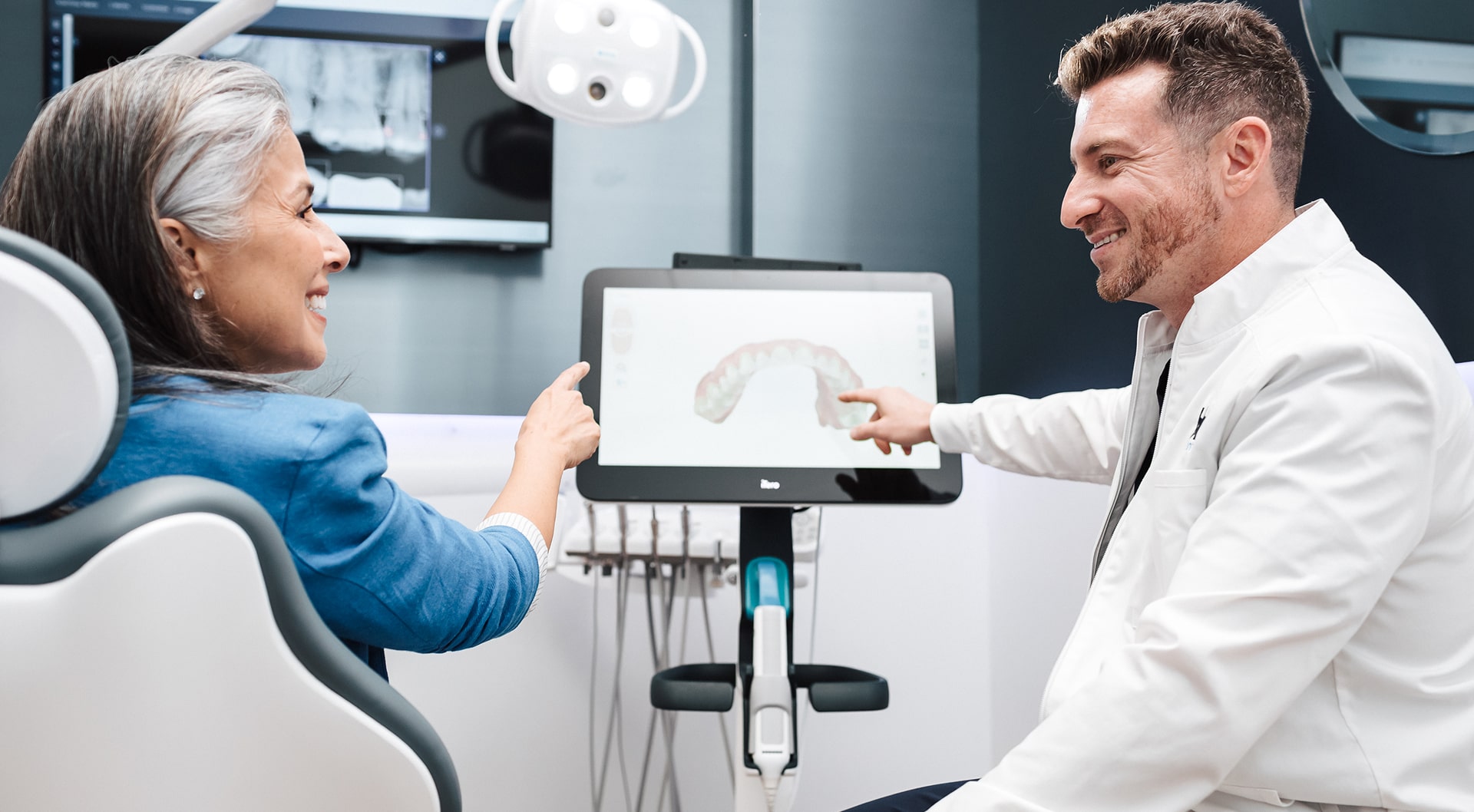 Dentist is smiling with a patient in a dental chair and pointing to the screen